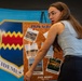 A young woman with glasses and long brown hair is presenting or pointing to a cork bulletin board displaying photos and information about a '2026 Static' display event. The board features photographs of various aircraft including an EA-37B, Dragonfly, T-38 Talon, and PT-17 Stearman, along with a reference to 'Omaha PD.' Behind her is a large blue banner featuring a military unit shield with the motto 'VIDEMUS.'