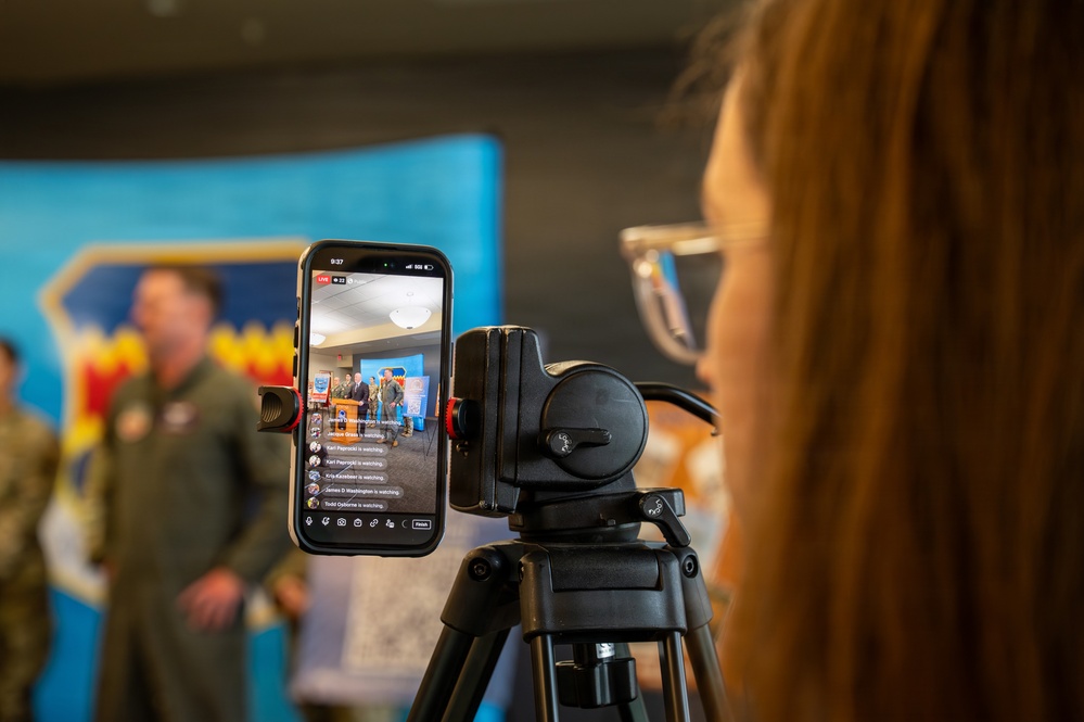 A person with red/auburn hair is managing a live social media stream on a smartphone mounted on a tripod. The phone screen shows an active live broadcast of what appears to be a military press conference or official event, with viewer names scrolling on screen. In the background, individuals in military flight suits are visible near a blue banner.
