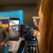 A person with red/auburn hair is managing a live social media stream on a smartphone mounted on a tripod. The phone screen shows an active live broadcast of what appears to be a military press conference or official event, with viewer names scrolling on screen. In the background, individuals in military flight suits are visible near a blue banner.