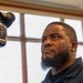 A man with a short beard and small earring is positioned beside a professional video camera mounted on a tripod, appearing to monitor or operate it. He is looking upward and to the side with a focused expression. The setting appears to be an indoor room with large windows providing natural light.