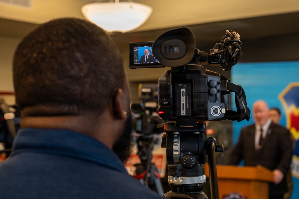 A videographer is filming a press conference or official briefing, with a man in a dark suit speaking at a podium in the background. The camera's flip-out LCD monitor displays a close-up of the speaker. A blue backdrop with what appears to be an official emblem or insignia is partially visible behind the speaker.