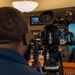 A videographer is filming a press conference or official briefing, with a man in a dark suit speaking at a podium in the background. The camera's flip-out LCD monitor displays a close-up of the speaker. A blue backdrop with what appears to be an official emblem or insignia is partially visible behind the speaker.