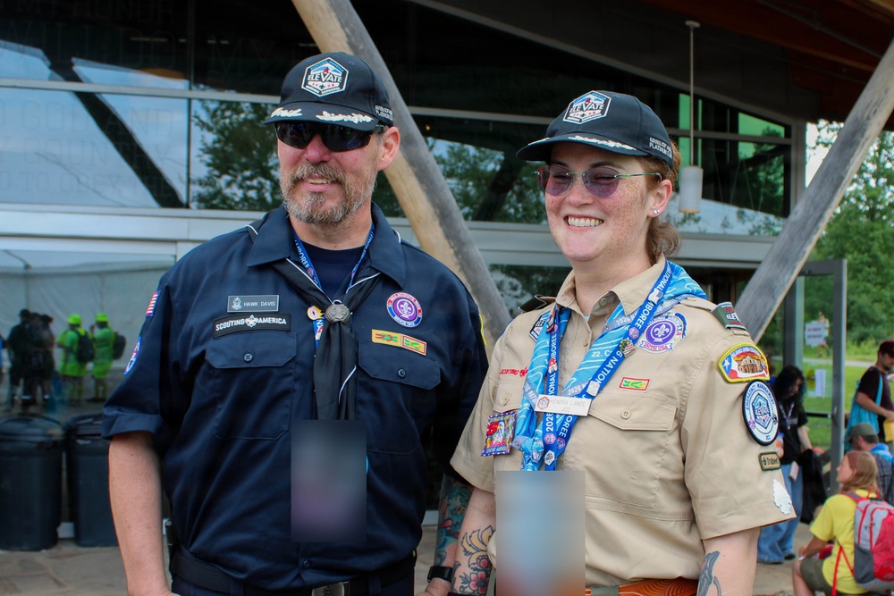 Two individuals wearing Scouting America uniforms and matching 'Elevate' caps pose together outdoors at what appears to be a large scouting event or jamboree. The person on the left wears a dark navy uniform with a name tag reading 'Hawk Davis' and a neckerchief, while the person on the right wears the traditional tan scout uniform adorned with multiple patches and event lanyards. Both are smiling and appear to be staff or adult leaders at the event.