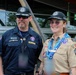 Two individuals wearing Scouting America uniforms and matching 'Elevate' caps pose together outdoors at what appears to be a large scouting event or jamboree. The person on the left wears a dark navy uniform with a name tag reading 'Hawk Davis' and a neckerchief, while the person on the right wears the traditional tan scout uniform adorned with multiple patches and event lanyards. Both are smiling and appear to be staff or adult leaders at the event.