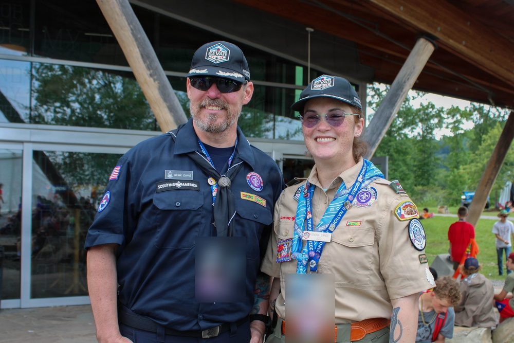 Two individuals wearing Scouting America uniforms pose together for a photo at what appears to be an outdoor scouting event. The person on the left wears a dark navy uniform with a name tag reading 'Hawk Davis' and a 'Scouting America' patch, while the person on the right wears the traditional tan scout uniform adorned with multiple patches and a blue neckerchief. Both wear matching 'Elevate' caps, suggesting this may be a specific scouting event or jamboree.