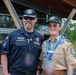Two individuals wearing Scouting America uniforms pose together for a photo at what appears to be an outdoor scouting event. The person on the left wears a dark navy uniform with a name tag reading 'Hawk Davis' and a 'Scouting America' patch, while the person on the right wears the traditional tan scout uniform adorned with multiple patches and a blue neckerchief. Both wear matching 'Elevate' caps, suggesting this may be a specific scouting event or jamboree.