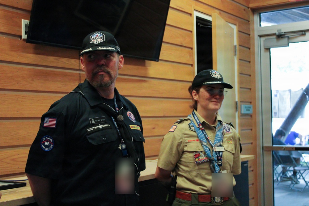 Two individuals wearing Scouting America uniforms are standing inside a wooden-paneled building. The person on the left wears a black uniform shirt with patches including a US flag and 'Scouting America' text, while the person on the right wears the traditional tan Scout uniform with a neckerchief and multiple badges. Both wear matching black caps with what appears to be an 'Elevate' logo.