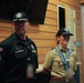 Two individuals wearing Scouting America uniforms are standing inside a wooden-paneled building. The person on the left wears a black uniform shirt with patches including a US flag and 'Scouting America' text, while the person on the right wears the traditional tan Scout uniform with a neckerchief and multiple badges. Both wear matching black caps with what appears to be an 'Elevate' logo.