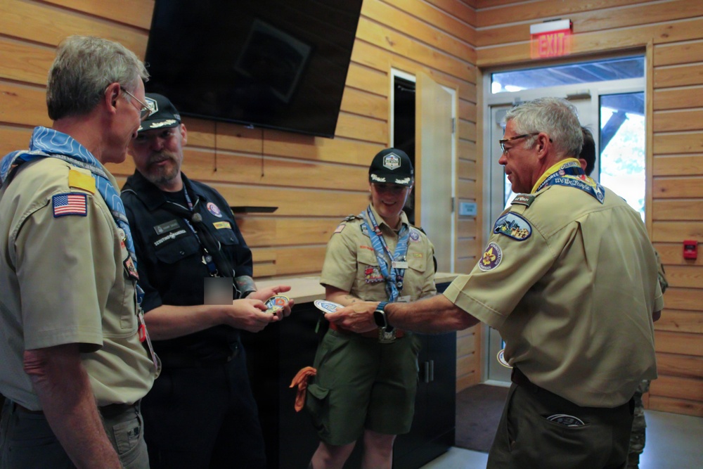 A group of four adults in Boy Scouts of America uniforms are gathered indoors in a wood-paneled room, engaged in what appears to be a patch or award exchange ceremony. Two individuals in tan BSA uniforms are exchanging or presenting circular patches/coins with another person, while a third person in a dark uniform observes. The setting appears to be a scout camp or lodge facility.