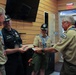 A group of four adults in Boy Scouts of America uniforms are gathered indoors in a wood-paneled room, engaged in what appears to be a patch or award exchange ceremony. Two individuals in tan BSA uniforms are exchanging or presenting circular patches/coins with another person, while a third person in a dark uniform observes. The setting appears to be a scout camp or lodge facility.