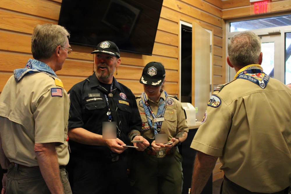 Four adults in Scouting America uniforms are gathered indoors, appearing to be in conversation and exchanging or reviewing documents. Two individuals wear dark navy uniforms with matching caps bearing a hexagonal emblem, while two others wear the traditional tan BSA uniform with neckerchiefs and patches. The setting appears to be a scout facility or lodge with wood-paneled walls.