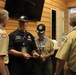 Four adults in Scouting America uniforms are gathered indoors, appearing to be in conversation and exchanging or reviewing documents. Two individuals wear dark navy uniforms with matching caps bearing a hexagonal emblem, while two others wear the traditional tan BSA uniform with neckerchiefs and patches. The setting appears to be a scout facility or lodge with wood-paneled walls.