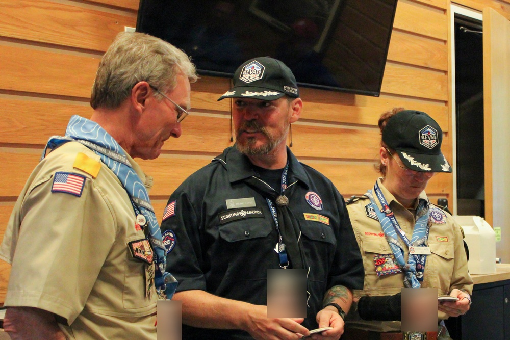 Three adults in Scouting America uniforms are gathered indoors, appearing to be at an official scouting event or ceremony. Two individuals wear tan BSA uniforms with patches, neckerchiefs, and lanyards, while the central figure wears a dark navy Scouting America staff shirt. All three wear matching dark caps with a hexagonal 'Elevate' logo, suggesting a specific scouting event or jamboree.