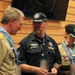 Three adults in Scouting America uniforms are gathered indoors, appearing to be at an official scouting event or ceremony. Two individuals wear tan BSA uniforms with patches, neckerchiefs, and lanyards, while the central figure wears a dark navy Scouting America staff shirt. All three wear matching dark caps with a hexagonal 'Elevate' logo, suggesting a specific scouting event or jamboree.