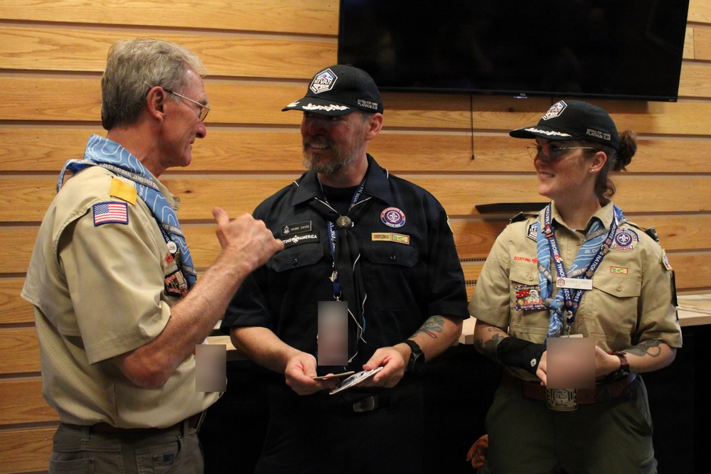 Three adults in Boy Scouts of America uniforms are engaged in conversation at what appears to be an indoor scouting event or gathering. Two individuals wear dark navy shirts with matching black caps, while one wears the traditional tan BSA uniform with a decorative neckerchief. They appear to be exchanging or reviewing items, possibly patches, cards, or awards.