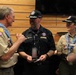 Three adults in Boy Scouts of America uniforms are engaged in conversation at what appears to be an indoor scouting event or gathering. Two individuals wear dark navy shirts with matching black caps, while one wears the traditional tan BSA uniform with a decorative neckerchief. They appear to be exchanging or reviewing items, possibly patches, cards, or awards.