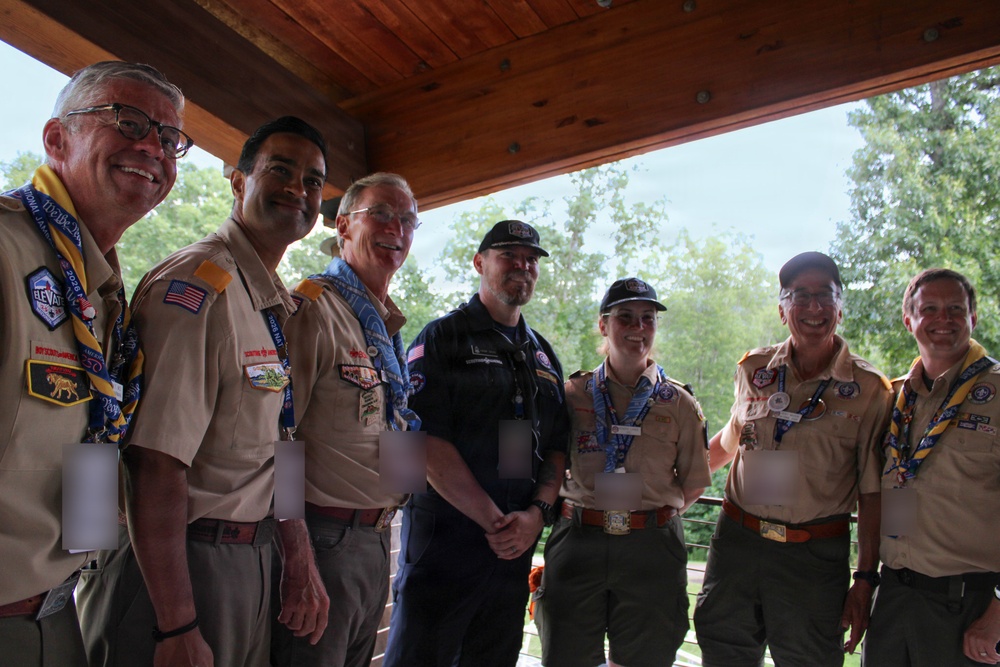 A group of seven adults posing together under a wooden pavilion or covered outdoor structure. Most are wearing Boy Scouts of America uniforms with patches, badges, and neckerchiefs, while one individual in the center is dressed in dark civilian clothing. The setting appears to be an outdoor scouting event or gathering, likely during summer based on the lush green trees visible in the background.