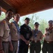 A group of seven adults posing together under a wooden pavilion or covered outdoor structure. Most are wearing Boy Scouts of America uniforms with patches, badges, and neckerchiefs, while one individual in the center is dressed in dark civilian clothing. The setting appears to be an outdoor scouting event or gathering, likely during summer based on the lush green trees visible in the background.