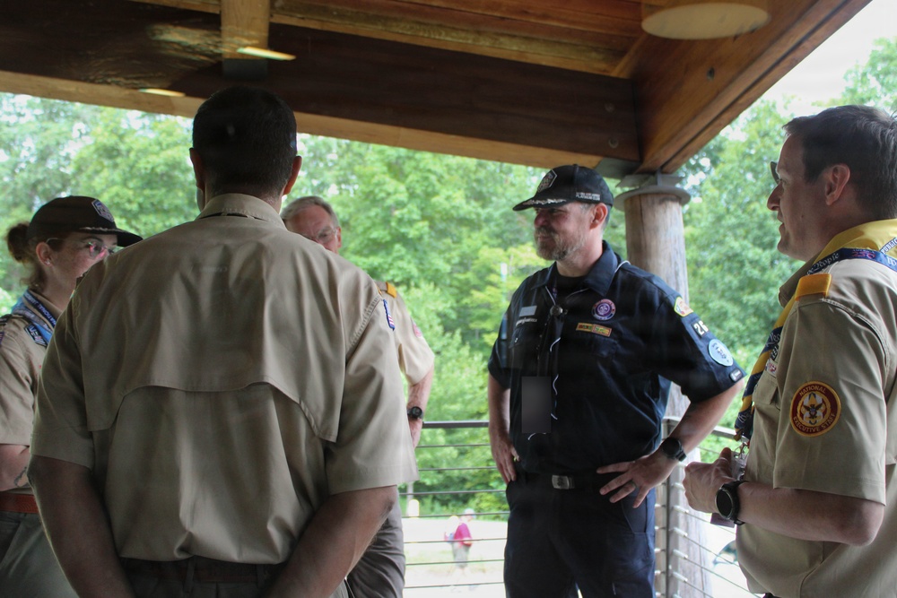 A group of individuals in scouting uniforms are gathered under a covered outdoor pavilion or deck, engaged in conversation. The group includes adults wearing tan Boy Scouts of America uniforms and one person in a dark navy uniform with a cap. The setting appears to be a wooded or camp environment based on the lush green trees visible in the background.