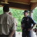 A group of individuals in scouting uniforms are gathered under a covered outdoor pavilion or deck, engaged in conversation. The group includes adults wearing tan Boy Scouts of America uniforms and one person in a dark navy uniform with a cap. The setting appears to be a wooded or camp environment based on the lush green trees visible in the background.