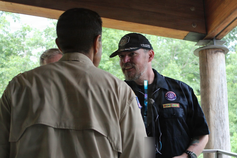 Three adults are gathered under a wooden pavilion or covered outdoor structure, engaged in conversation. The man facing the camera wears a dark navy shirt with Scout-related patches and a black cap with 'National Scout Jamboree Platinum Club' text. The setting appears to be a scouting event or camp, with lush green trees visible in the background.