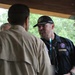 Three adults are gathered under a wooden pavilion or covered outdoor structure, engaged in conversation. The man facing the camera wears a dark navy shirt with Scout-related patches and a black cap with 'National Scout Jamboree Platinum Club' text. The setting appears to be a scouting event or camp, with lush green trees visible in the background.