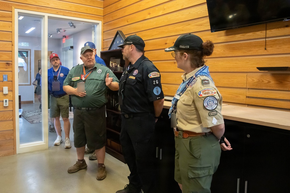 A group of people in various uniforms and casual attire are gathered inside a wood-paneled room, engaged in conversation. Three individuals in the foreground appear to be in scouting or organizational uniforms with patches and badges, while two others are visible through a glass door in the background. The setting appears to be an indoor facility or visitor center.