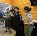A group of people in various uniforms and casual attire are gathered inside a wood-paneled room, engaged in conversation. Three individuals in the foreground appear to be in scouting or organizational uniforms with patches and badges, while two others are visible through a glass door in the background. The setting appears to be an indoor facility or visitor center.