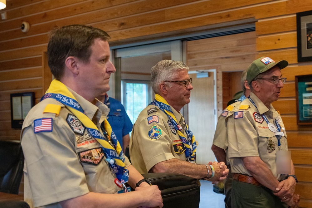 Three adult men in Boy Scouts of America uniforms stand attentively in what appears to be a lodge or meeting hall with wood-paneled walls. They are wearing tan scout shirts adorned with numerous patches and badges, along with yellow and blue 'We The People' neckerchiefs/lanyards. A fourth person in a blue shirt is partially visible in the background.