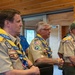 Three adult men in Boy Scouts of America uniforms stand attentively in what appears to be a lodge or meeting hall with wood-paneled walls. They are wearing tan scout shirts adorned with numerous patches and badges, along with yellow and blue 'We The People' neckerchiefs/lanyards. A fourth person in a blue shirt is partially visible in the background.