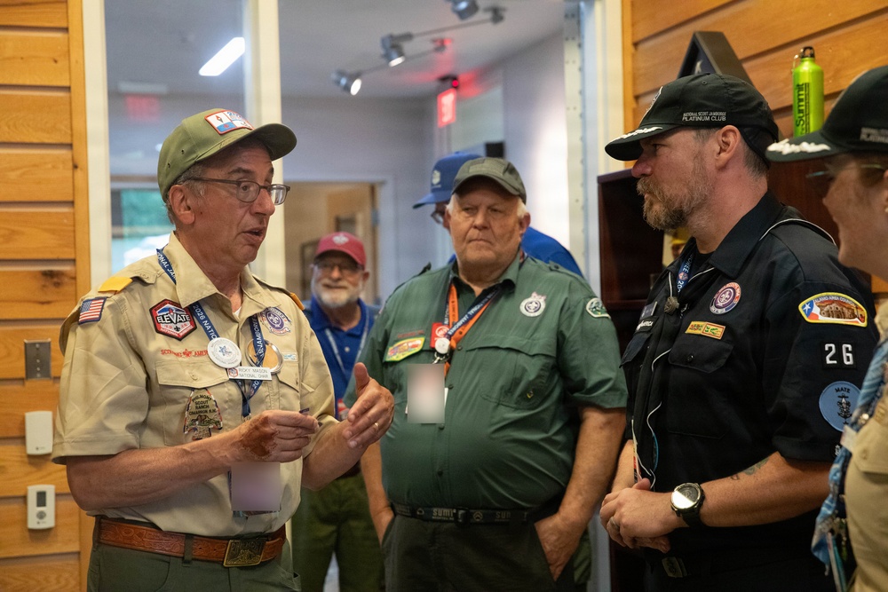 A group of adults in scouting uniforms and attire are gathered indoors, engaged in conversation. The man on the left, wearing a tan BSA (Boy Scouts of America) uniform with numerous patches and badges, appears to be speaking to others in the group. The setting appears to be a scouting facility or event venue with wood-paneled walls.