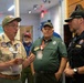 A group of adults in scouting uniforms and attire are gathered indoors, engaged in conversation. The man on the left, wearing a tan BSA (Boy Scouts of America) uniform with numerous patches and badges, appears to be speaking to others in the group. The setting appears to be a scouting facility or event venue with wood-paneled walls.