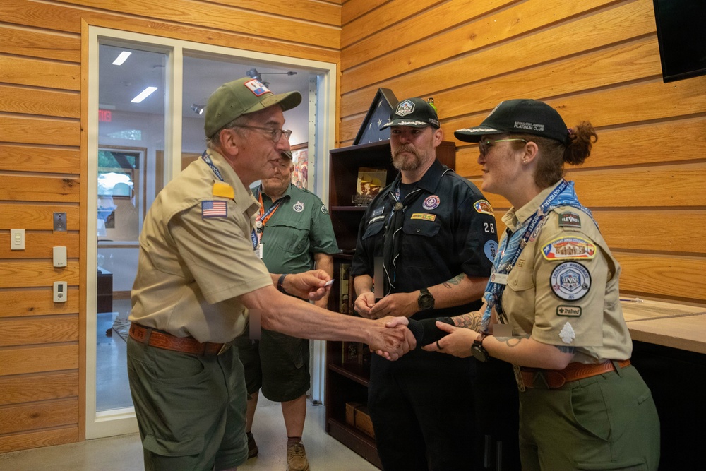 Four individuals in scouting uniforms are gathered in a wood-paneled room, with two of them engaged in a handshake. The participants are wearing various scout-related patches, lanyards, and caps, suggesting an official recognition or ceremonial exchange. The setting appears to be an indoor scout facility or administrative office.