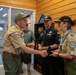 Four individuals in scouting uniforms are gathered in a wood-paneled room, with two of them engaged in a handshake. The participants are wearing various scout-related patches, lanyards, and caps, suggesting an official recognition or ceremonial exchange. The setting appears to be an indoor scout facility or administrative office.