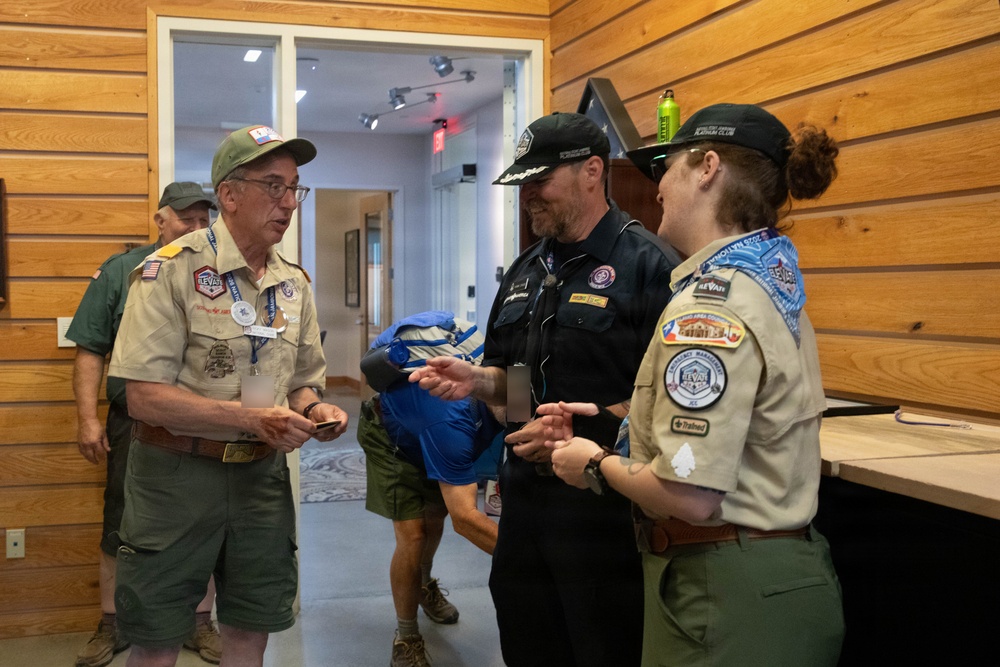 Three adults are engaged in conversation inside a wood-paneled building, with two wearing Boy Scouts of America uniforms adorned with patches and badges. A third person in a black uniform and cap appears to be showing or exchanging something with the others. A fourth individual is visible in the background near a doorway.