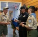 Three adults are engaged in conversation inside a wood-paneled building, with two wearing Boy Scouts of America uniforms adorned with patches and badges. A third person in a black uniform and cap appears to be showing or exchanging something with the others. A fourth individual is visible in the background near a doorway.