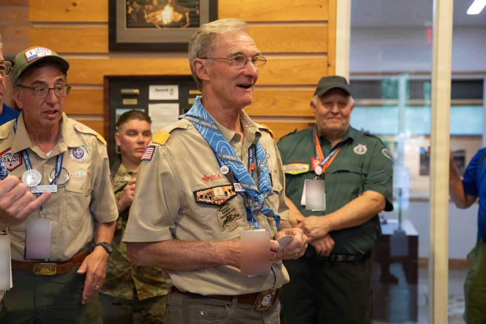 A group of Boy Scouts of America (BSA) adult leaders and volunteers are gathered indoors, likely at a National Jamboree event based on visible neckerchief and patches. The central figure, an older man wearing a tan BSA uniform with multiple patches and a blue National Jamboree neckerchief, appears to be speaking or presenting. The setting appears to be an indoor facility with wood-paneled walls and framed displays.