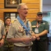 A group of Boy Scouts of America (BSA) adult leaders and volunteers are gathered indoors, likely at a National Jamboree event based on visible neckerchief and patches. The central figure, an older man wearing a tan BSA uniform with multiple patches and a blue National Jamboree neckerchief, appears to be speaking or presenting. The setting appears to be an indoor facility with wood-paneled walls and framed displays.