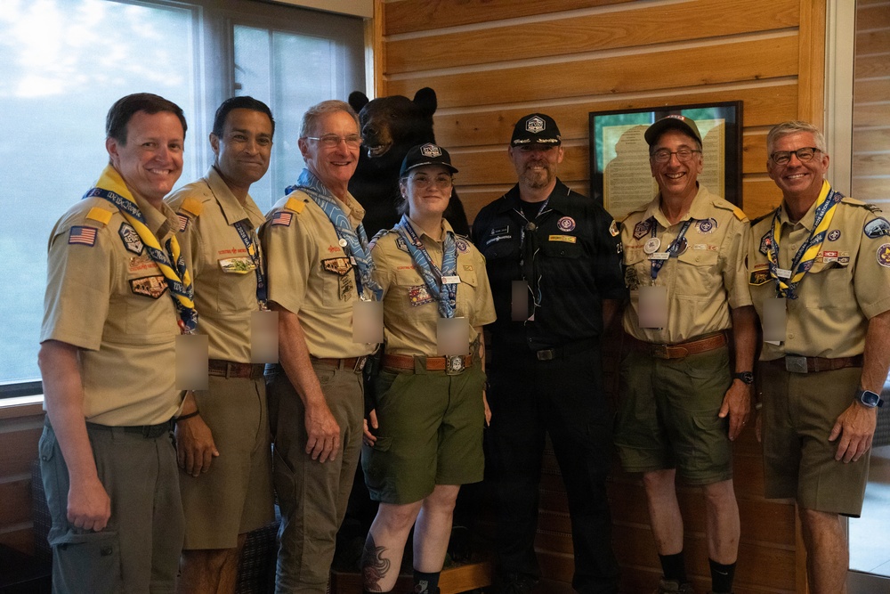 A group of seven individuals pose together indoors, most wearing Boy Scouts of America uniforms with patches, neckerchiefs, and merit badges. The setting appears to be a lodge or scout facility with wood-paneled walls, a framed document, and a taxidermied black bear visible in the background. The group appears to be at a scouting event or gathering.
