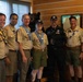A group of seven individuals pose together indoors, most wearing Boy Scouts of America uniforms with patches, neckerchiefs, and merit badges. The setting appears to be a lodge or scout facility with wood-paneled walls, a framed document, and a taxidermied black bear visible in the background. The group appears to be at a scouting event or gathering.
