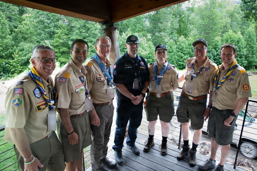 A group of seven adults posed together under a covered outdoor pavilion or deck, most wearing Boy Scouts of America uniforms with patches, neckerchiefs, and lanyards. One individual in the center is dressed in dark tactical/staff clothing, contrasting with the tan BSA uniforms worn by the others. The setting appears to be a wooded scout camp or outdoor facility during daytime.