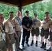 A group of seven adults posed together under a covered outdoor pavilion or deck, most wearing Boy Scouts of America uniforms with patches, neckerchiefs, and lanyards. One individual in the center is dressed in dark tactical/staff clothing, contrasting with the tan BSA uniforms worn by the others. The setting appears to be a wooded scout camp or outdoor facility during daytime.