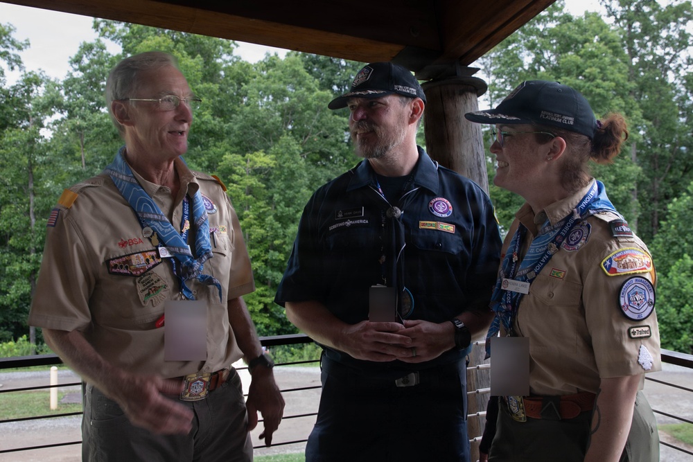 Three adults in Boy Scouts of America (BSA) uniforms are engaged in conversation under a covered outdoor pavilion. Two individuals wear tan BSA uniforms adorned with numerous patches and neckerchiefs, while the third wears a dark navy BSA uniform. They appear to be at a scouting event or jamboree, based on their lanyards, badges, and insignia.