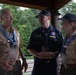 Three adults in Boy Scouts of America (BSA) uniforms are engaged in conversation under a covered outdoor pavilion. Two individuals wear tan BSA uniforms adorned with numerous patches and neckerchiefs, while the third wears a dark navy BSA uniform. They appear to be at a scouting event or jamboree, based on their lanyards, badges, and insignia.