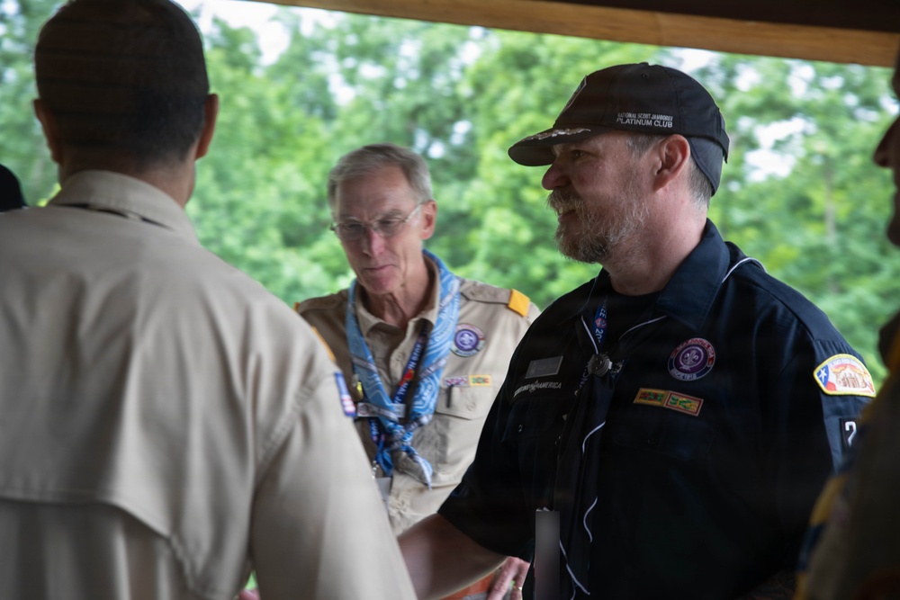 Three men are gathered under a covered outdoor structure, appearing to be engaged in conversation or a greeting. Two of the men are wearing Boy Scouts of America uniforms, while the third wears a dark navy uniform with patches and a cap reading 'National Scout Jamboree Platinum Club'. The setting appears to be an outdoor scouting event surrounded by green trees.