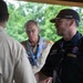Three men are gathered under a covered outdoor structure, appearing to be engaged in conversation or a greeting. Two of the men are wearing Boy Scouts of America uniforms, while the third wears a dark navy uniform with patches and a cap reading 'National Scout Jamboree Platinum Club'. The setting appears to be an outdoor scouting event surrounded by green trees.