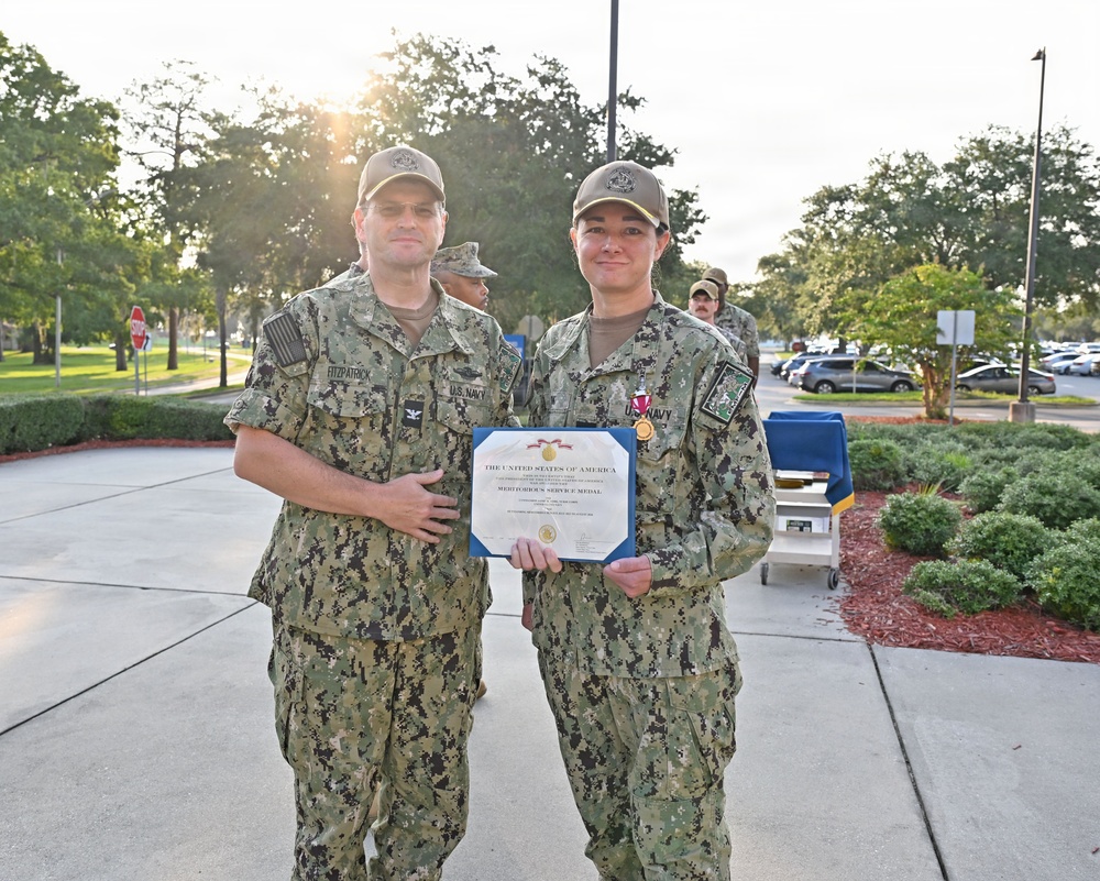 Two U.S. Navy personnel in NWU Type III (woodland camouflage) uniforms pose together holding a Meritorious Service Medal certificate. The service member on the right is wearing the Meritorious Service Medal on their uniform and holding the accompanying certificate. The ceremony appears to be taking place outdoors on a military installation.