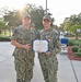 Two U.S. Navy personnel in NWU Type III (woodland camouflage) uniforms pose together holding a Meritorious Service Medal certificate. The service member on the right is wearing the Meritorious Service Medal on their uniform and holding the accompanying certificate. The ceremony appears to be taking place outdoors on a military installation.
