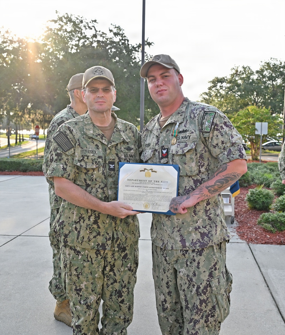 Two U.S. Navy personnel in NWU Type III (woodland digital camouflage) uniforms pose together holding a Department of the Navy certificate for the Navy and Marine Corps Achievement Medal. The individual on the left, identified by name tape as 'FITZPATRICK,' appears to be a senior officer presenting the award to the recipient on the right, identified as 'SOUSA,' who is wearing the medal on his uniform. The ceremony appears to be taking place outdoors on a military installation.