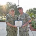 Two U.S. Navy personnel in NWU Type III (woodland digital camouflage) uniforms pose together holding a Department of the Navy certificate for the Navy and Marine Corps Achievement Medal. The individual on the left, identified by name tape as 'FITZPATRICK,' appears to be a senior officer presenting the award to the recipient on the right, identified as 'SOUSA,' who is wearing the medal on his uniform. The ceremony appears to be taking place outdoors on a military installation.